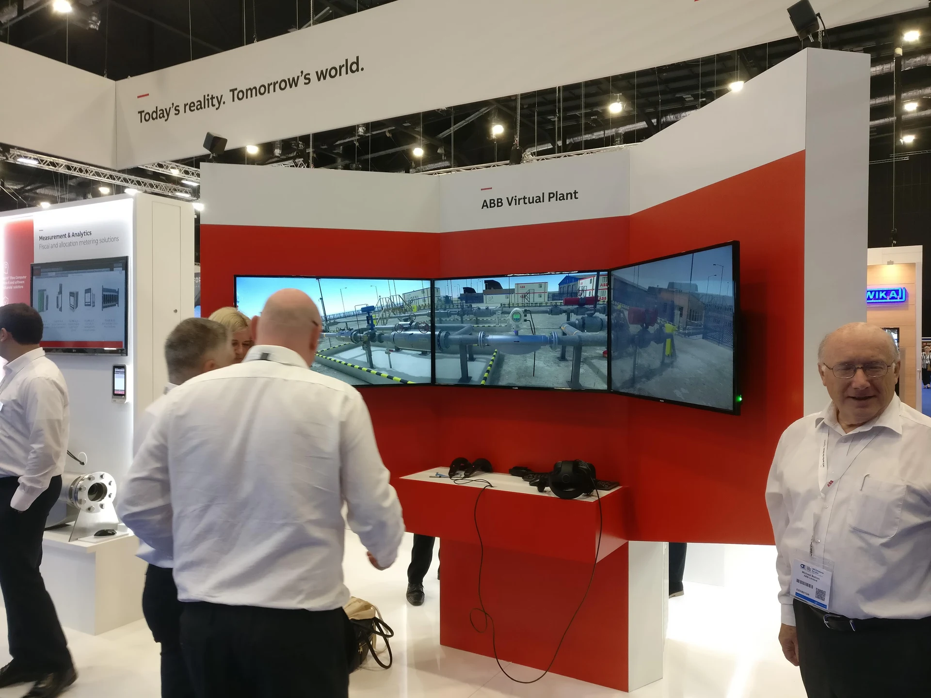 ABB Virtual Plant exhibition stand with three angled screens displaying the simulated plant and an HTC Vive headset on the counter.