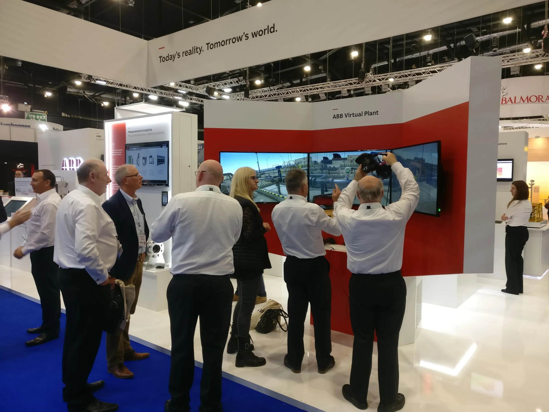 Visitors gathered at the ABB Virtual Plant stand as a participant lifts the VR headset in front of the triple-screen display.