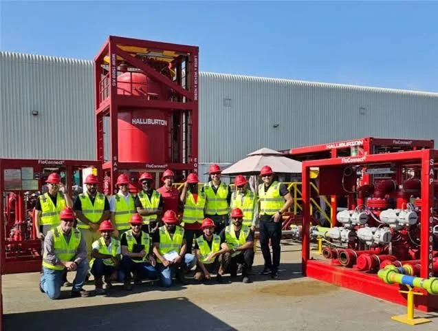 A group wearing hard hats and high-visibility vests stands beside red Halliburton FloConnect equipment skids.